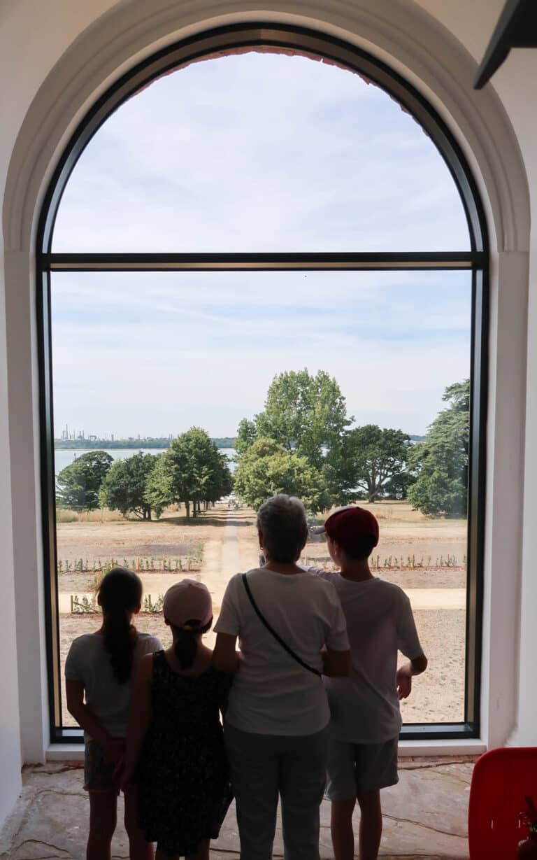 Four people—an adult and three children—stand inside, silhouetted against a large arched window, looking out at a garden path, trees, and water under a bright sky in this serene Weekend In Pictures moment.