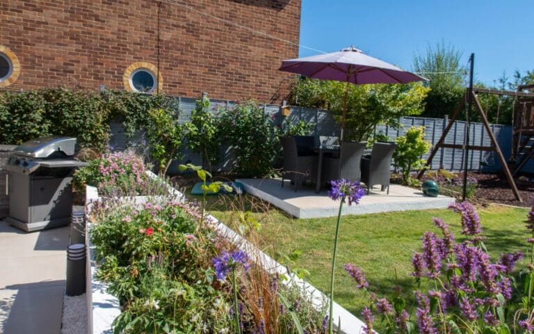 Enjoy a Family Garden Tour in this sunny back garden featuring colourful flowers, a patio with table and chairs under a purple umbrella, a barbecue, and a swing set by the brick wall and wooden fence.
