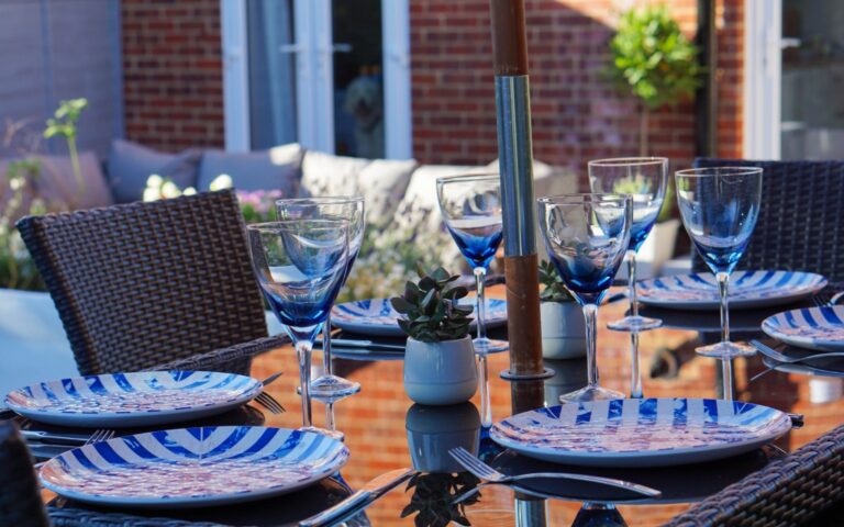 A patio dining table set for six, perfect for a Family Garden Tour, features blue and white striped plates, blue-rimmed wine glasses, cutlery, and small potted succulents, with wicker chairs and a brick house in the background.