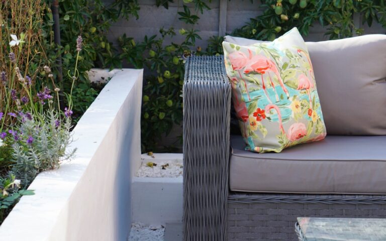 A grey wicker outdoor sofa with light grey cushions and a colourful pillow featuring pink flamingos and green foliage, perfect for relaxing during a Family Garden Tour, set on a patio next to plants and a glass-topped table.