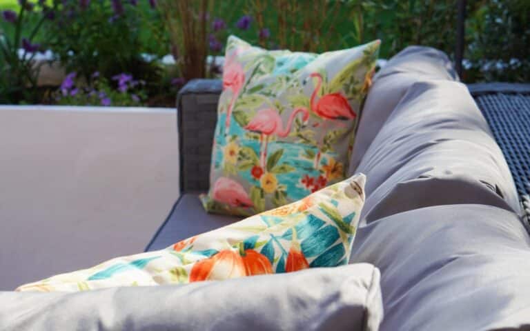 A close-up of a grey outdoor sofa with two colourful decorative cushions featuring tropical patterns, perfect for relaxing during a Family Garden Tour. Green plants and purple flowers create a vibrant, blurred background.