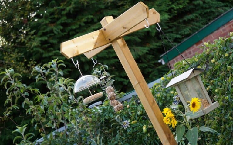 A wooden bird feeder pole in a garden holds various bird feeders and bird food. Sunflowers grow at the base, while green shrubs and trees create a lush background, making it easy to feed the birds with household scraps.
