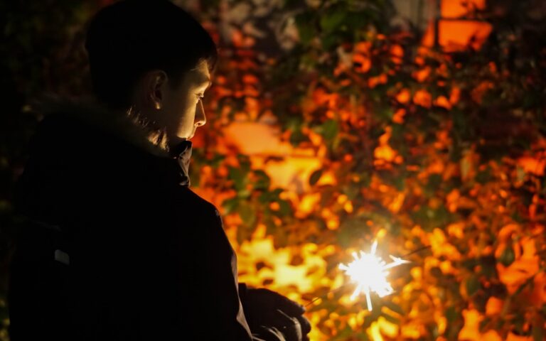 A person stands outdoors at night in winter, holding a lit sparkler. Warm orange light from the sparkler and nearby Philips Hue outdoor lighting illuminates their face, whilst the background features leafy plants in shadow.