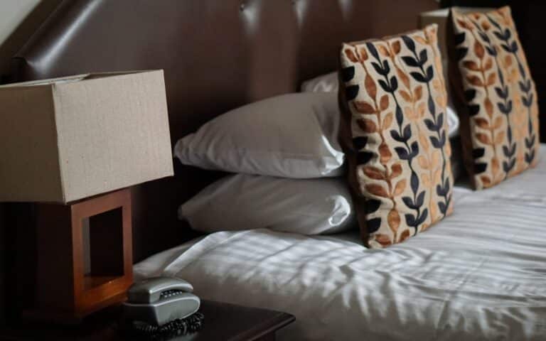 A neatly made bed with white linen, two leafy-patterned pillows, and two plain white pillows. A brown headboard, a lamp with a square shade, and a telephone are on the bedside table—ideal for a relaxing Red Letter Days experience.