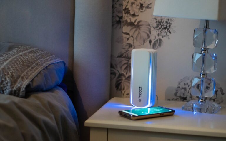 A Foobot Indoor Air Quality Monitor emits blue light onto a smartphone on a bedside table next to a bed with a grey duvet and a clear crystal lamp, helping to improve air quality in your bedroom.