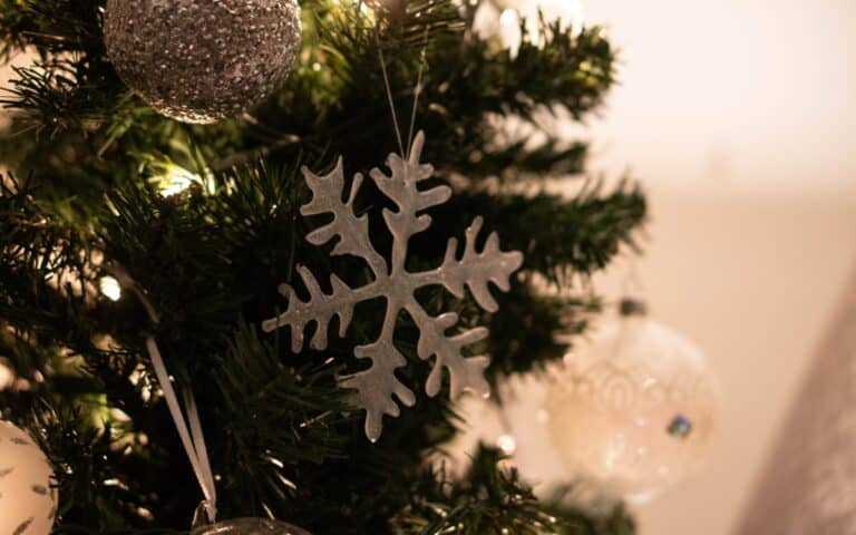 A close-up of a decorated Christmas tree with handmade snowflakes, a glittery silver bauble, and a white ornament hanging among green pine needles and soft white lights—perfect inspiration for DIY crafts with a glue gun.