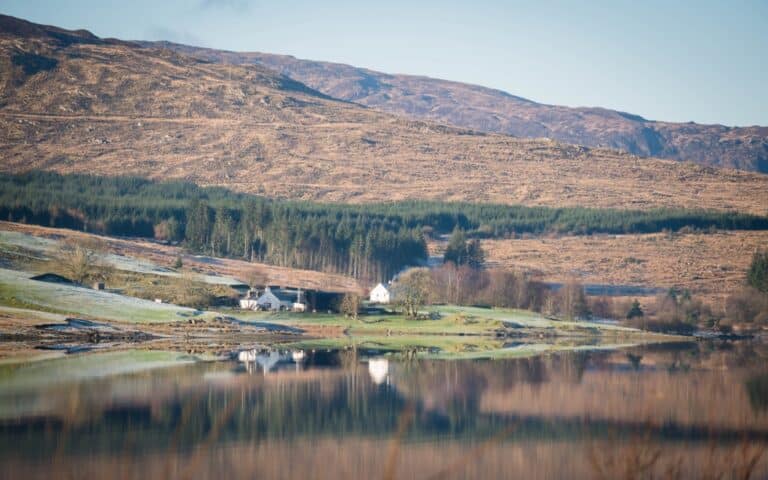 A white cottage sits among trees near the shore of a calm loch, with hills covered in brown grass and green forest beyond—capturing the serene beauty of Christmas in Scotland, all reflected in the water under a clear sky.