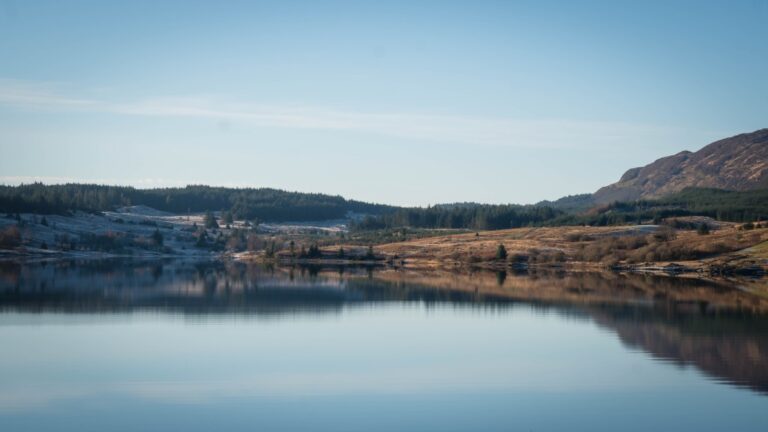 A calm lake reflects the surrounding hills and a clear blue sky, with forested and grassy areas in the background under natural daylight—a tranquil scene reminiscent of Christmas in Scotland.