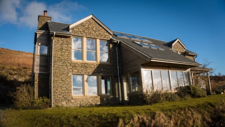 A modern house with stone and wood exterior, large windows reflecting the sky, and a sloped roof with solar panels sits on a grassy hill under a blue sky—evoking the cosy charm of Christmas in Scotland.