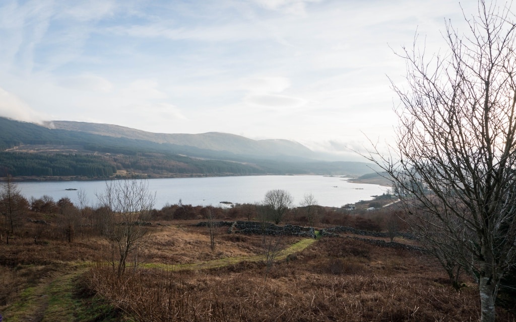 A scenic view of a calm lake surrounded by hills and sparse trees under a cloudy sky, with a winding path and dry grass in the foreground captures the peaceful charm of Christmas in Scotland.