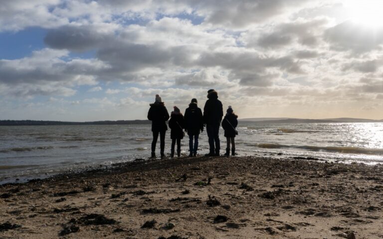 Me and Mine: In January, a group of five people, warmly dressed, stand together on a sandy beach facing the water under a cloudy sky, with sunlight reflecting on the sea.