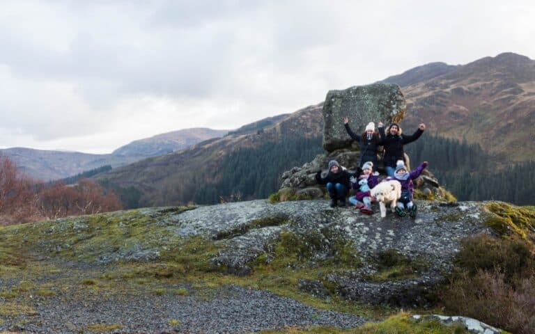 Me and Mine: A group of people and a dog pose cheerfully on a rocky hilltop, with mossy ground and mountains covered in trees in the background under a cloudy December sky.