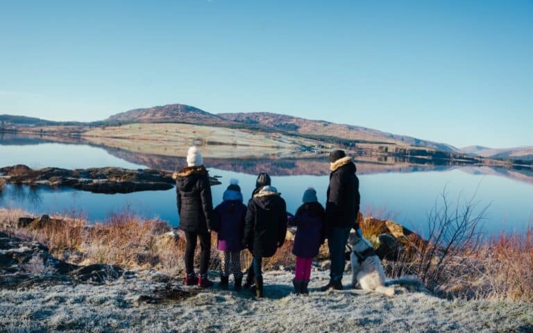 A family of four, dressed in winter clothing, stands with their dog by a calm lake in December, looking at distant hills under a clear blue sky. Frost covers the ground and the landscape reflects on the water for this special Me and Mine moment.