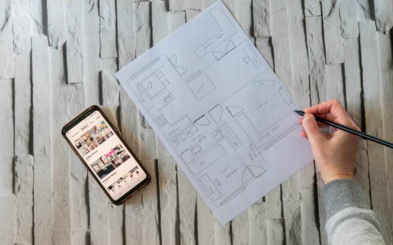 A person sketches a home extension on a floor plan with a pencil whilst holding the paper. Nearby, a mobile phone displays renovation images, all set on a light grey textured surface.