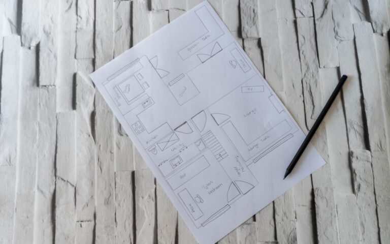 A hand-drawn floor plan on white paper with labelled rooms sits on a textured white surface next to a black pencil, illustrating ideas for home improvement or extension projects.
