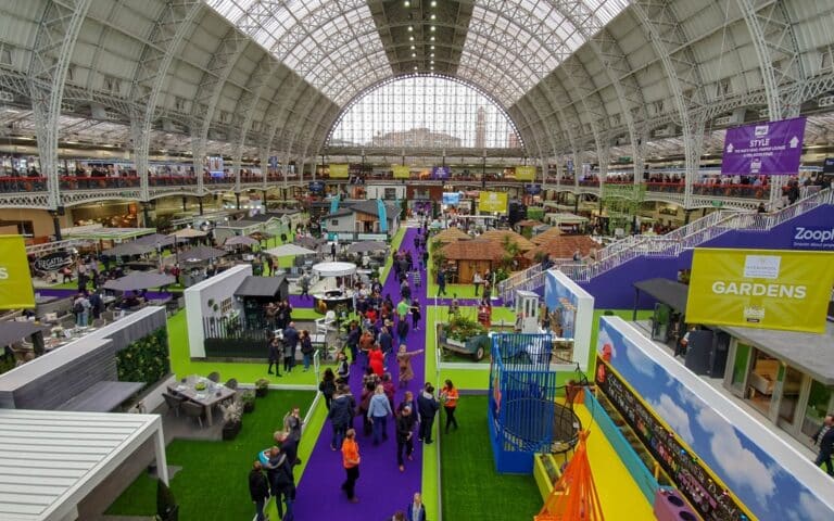 A large indoor exhibition hall at Olympia London with high arched glass ceilings, colourful stands, garden displays, and crowds walking on purple carpeted pathways. Signs indicate various themed areas at the Ideal Home Show, including “GARDENS.”.