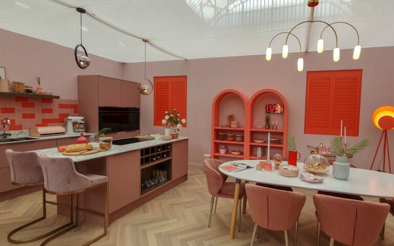 Modern kitchen and dining area with pink and coral decor, inspired by the Ideal Home Show at Olympia London. Features a central island, velvet chairs, open shelving, contemporary lighting, warm tones, and stylish accessories.