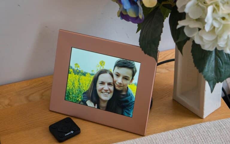 A digital photo frame on a wooden table displays a smiling woman and a boy in a field of yellow flowers—an ideal addition to any 2019 Father’s Day Gift Guide. Nearby are a remote control and a vase with white and purple flowers.