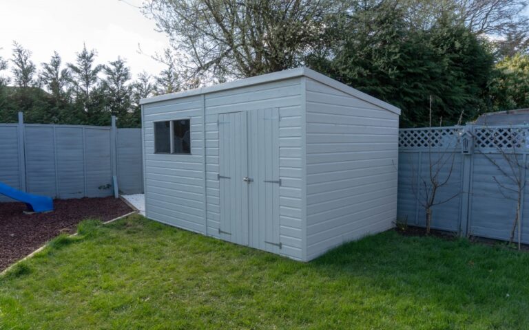 A light grey wooden garden shed with a window and double doors stands on green grass in a fenced back garden, set atop a durable Plastic Shed Base, surrounded by trees and plants.