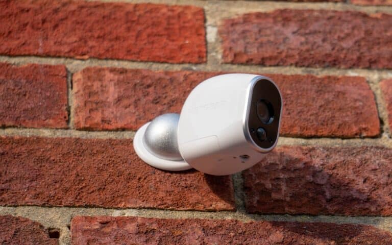 A white wireless security camera is mounted on a red brick wall, positioned at an angle to monitor the area. The camera has a rounded design, is attached to a metallic base, and offers features highlighted in many security camera reviews.