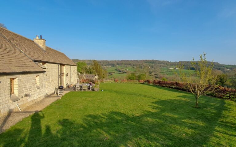 A wonderful stone house with a patio sits next to a large, green lawn overlooking a scenic countryside—perfect for an Easter holiday amid rolling hills and trees beneath a clear blue sky. A lone tree stands on the lawn near the edge.