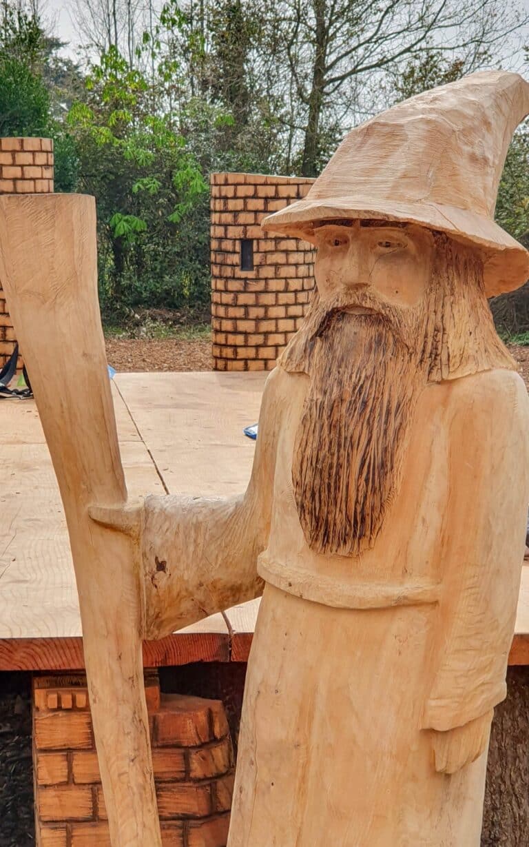 A wooden sculpture of a wizard with a long beard and pointed hat, holding a tall staff, stands on a platform outdoors in Royal Victoria Country Park, surrounded by trees and brick structures.