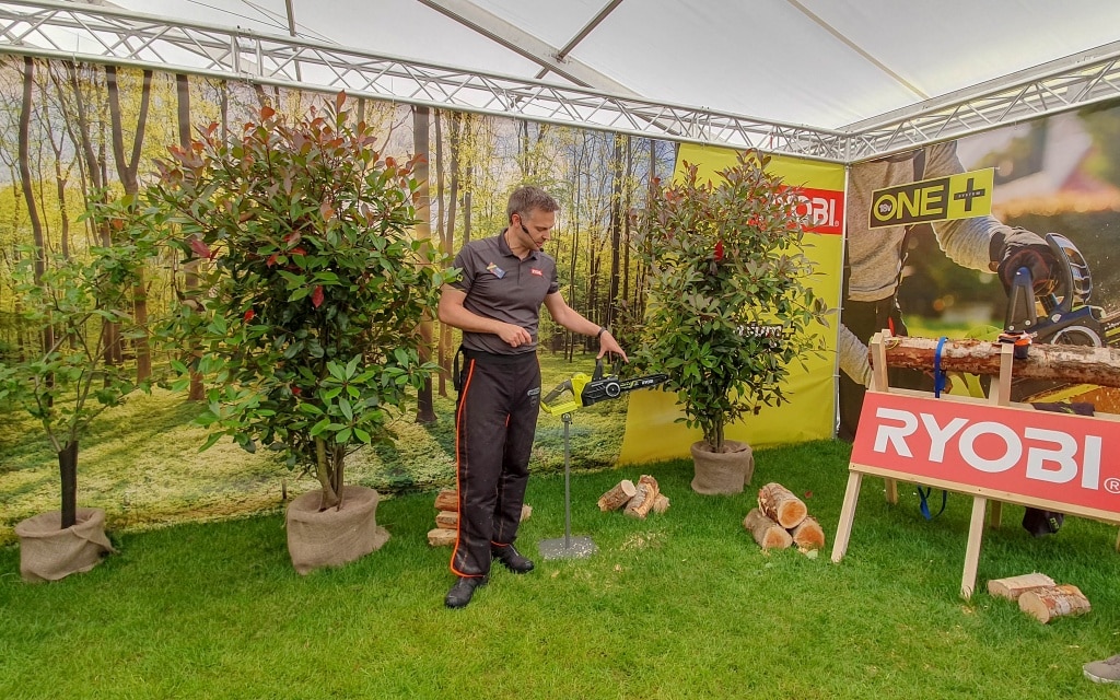A man demonstrates a Ryobi chainsaw tool under a white canopy at a conference, surrounded by potted shrubs and a Ryobi display table with wood logs, set against a scenic forest backdrop.