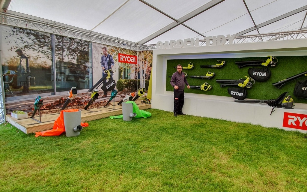 A man stands in a tented display area at the 2019 conference, surrounded by Ryobi garden tools—including a chainsaw—mounted on a wall and arranged on the floor, with a large GARDEN E sign and promotional banners on grass.