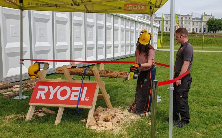 A woman in safety gear uses a Ryobi chainsaw to cut a log on a stand under a yellow Ryobi tent at the 2019 conference, whilst a man observes nearby. Wood chips cover the grass, with a large white structure in the background.