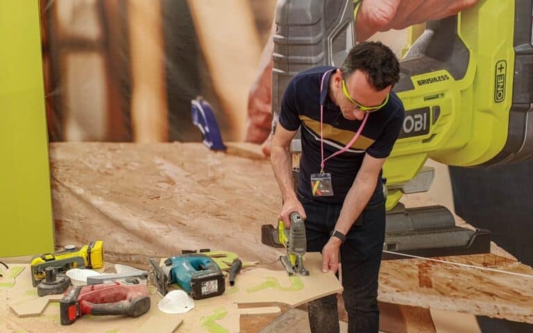 A man wearing safety glasses uses a Ryobi power tool to cut a piece of wood on a workbench, surrounded by other tools and sawdust.