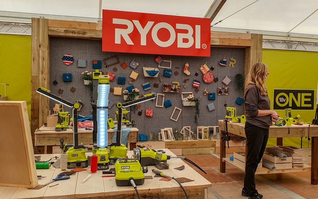 A woman stands in a well-lit workshop area with various Ryobi power tools, including a chainsaw, displayed on tables. A large red Ryobi sign hangs above a decorated wall showcasing crafts and wooden shapes from the 2019 Conference.
