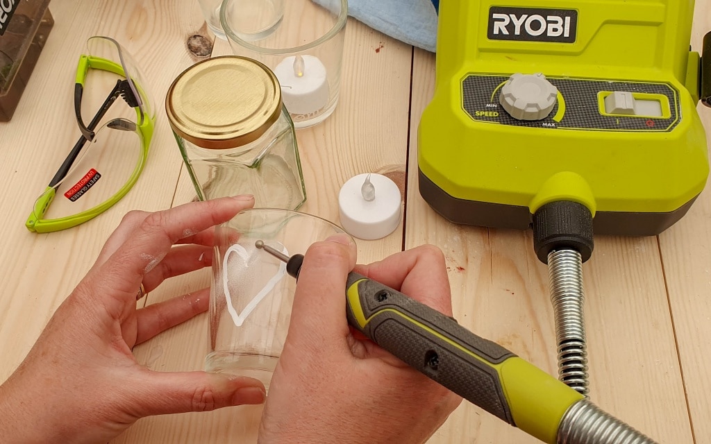 A person uses a Ryobi engraving tool to etch a heart shape onto a glass jar. Safety glasses, a Kilner jar, tea lights, and other crafting supplies from the 2019 Conference are on the wooden table.