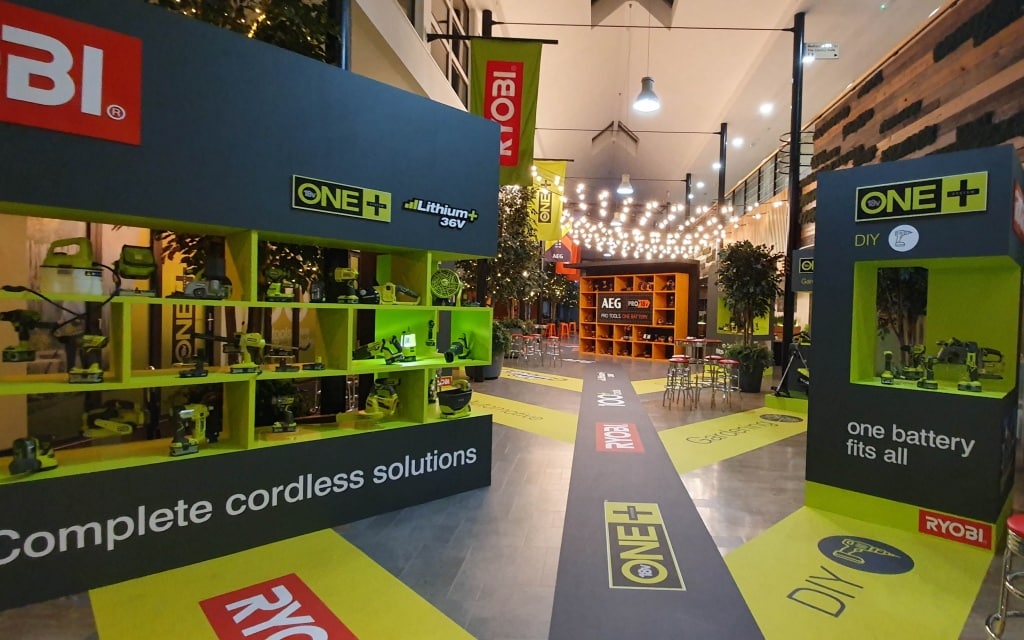 A showroom displays Ryobi ONE+ cordless power tools, including a chainsaw, on shelves with bright signage and banners. Fairy lights hang from the ceiling, lounge seating is in the background, and floor graphics highlight the 2019 Conference branding.