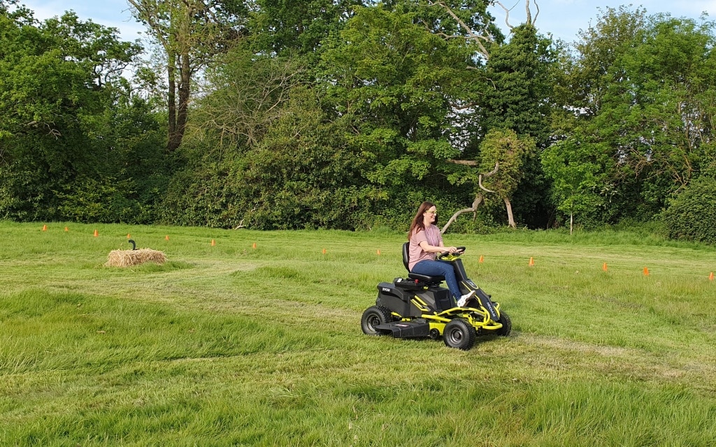 A woman in a pink shirt rides a yellow and black Ryobi lawnmower on a grassy field, with trees and a hay bale visible in the background.