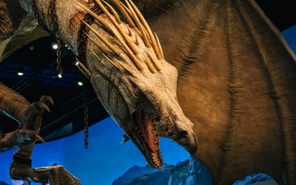 A close-up of a large, detailed dragon with beige, textured scales, spiky horns, and open mouth showing sharp teeth, set against a background of mountains and a blue sky at Warner Bros Studio Tour London.