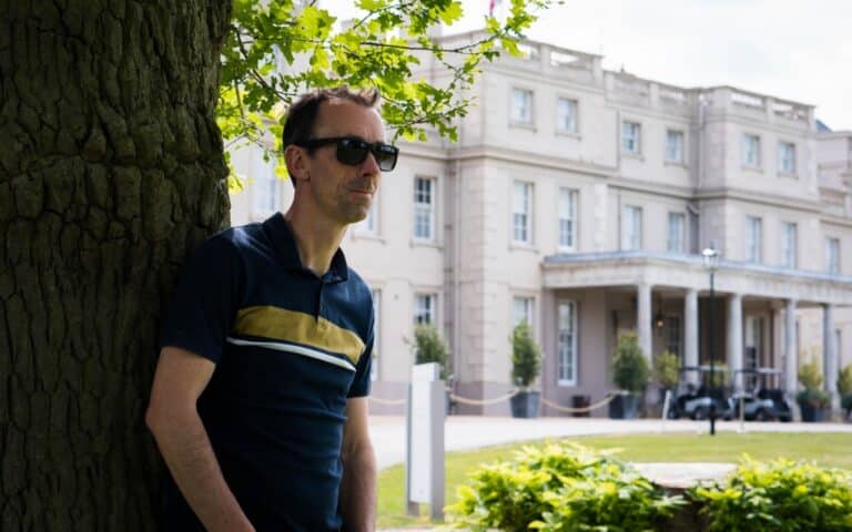 A man wearing sunglasses and a navy striped shirt leans against a tree in front of a large, elegant white building surrounded by greenery on a sunny day—perfect inspiration for your 2019 Father’s Day Gift Guide.