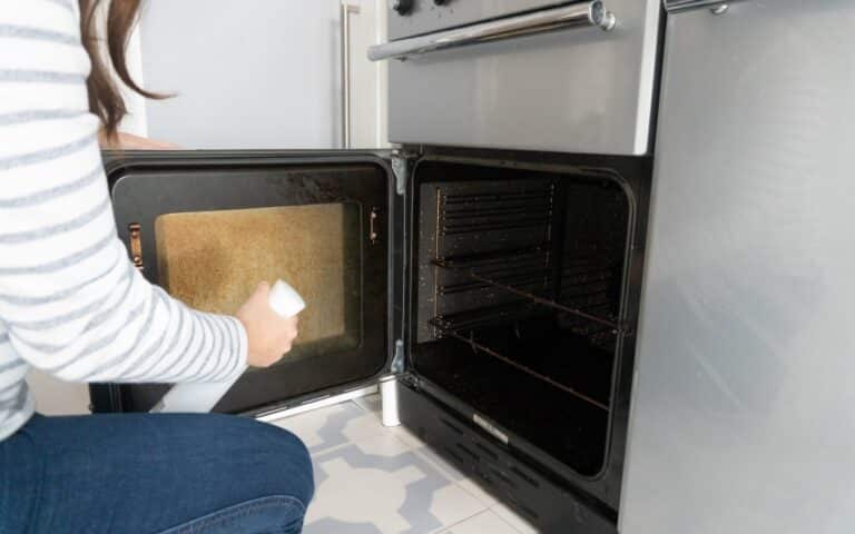 A person in a striped shirt is kneeling and spraying cleaner from the Koh Starter Kit on the open door of a dirty oven, choosing toxic-free cleaning in the kitchen.