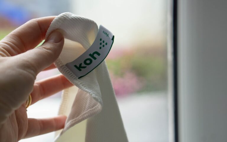A hand holds a white cleaning cloth labelled koh near a window, with a blurred outdoor view in the background—a glimpse into toxic-free cleaning with the Koh Starter Kit.