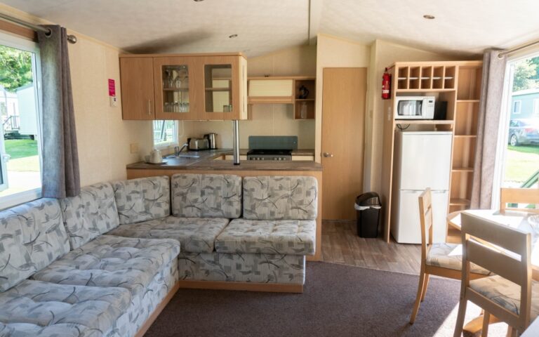 Modern mobile home interior with a patterned L-shaped sofa, small kitchen area, fridge, microwave, dining table with chairs, and large windows letting in natural light—perfect for a weekend away at Holywell Bay holiday park.