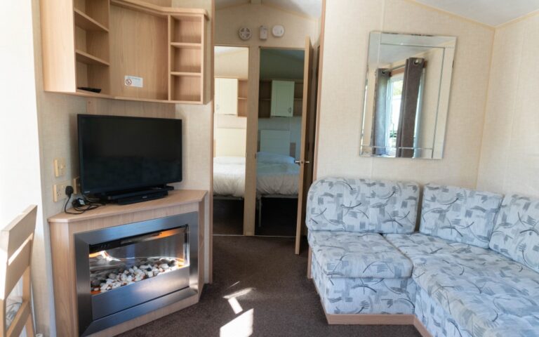 A cosy living room with a patterned light blue sofa, a flat-screen TV on a wooden stand above an electric fireplace, a large wall mirror, and views into a bedroom—perfect for a relaxing weekend away at Holywell Bay Holiday Park.