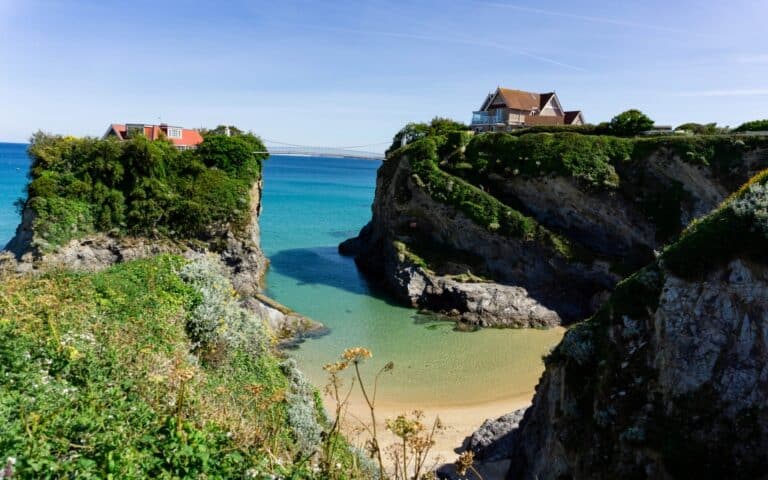 Two houses sit atop rocky cliffs overlooking the turquoise sea in Newquay, connected by a footbridge. Below, a sandy beach nestles between cliffs, with lush greenery and wildflowers—perfect for exploring Britain’s scenic coastline.