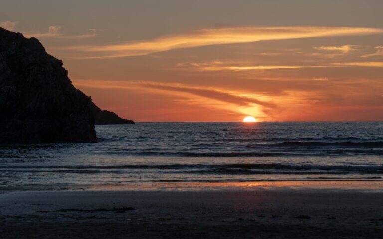 A golden-orange sunset over the ocean near Newquay, with the sun partially visible on the horizon. Gentle waves wash onto the sandy shore, and a rocky cliff is silhouetted on the left—perfect for exploring the Best of Britain’s coast.