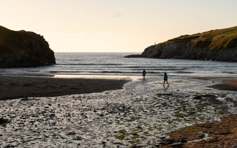 Two people walk along a sandy Newquay beach at low tide, with shallow water reflecting the sky. Rocky cliffs frame the beach on both sides, and the sea is calm under a soft, golden evening light—truly the Best of Britain.