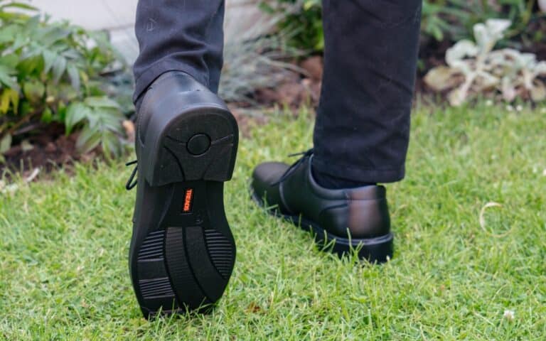 A person wearing hard-wearing school shoes and dark trousers is walking on green grass. The focus is on the shoe's sole, showing its distinctive treads and a small orange label.