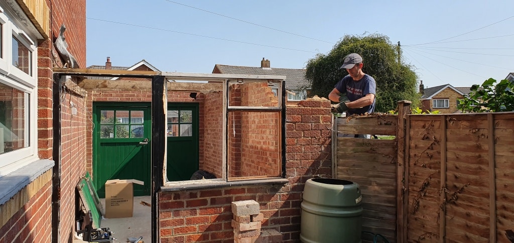 A man wearing a cap builds a brick wall next to a wooden fence in a back garden, possibly as part of an extension to the existing house. A green door and water butt are visible nearby under sunny skies.