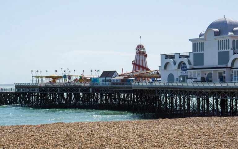 South Parade Pier in Southsea features a domed building, fairground rides, Kidz Island attractions, colourful flags, and a helter-skelter slide stretching over the water, with a pebble beach in the foreground beneath a clear sky.