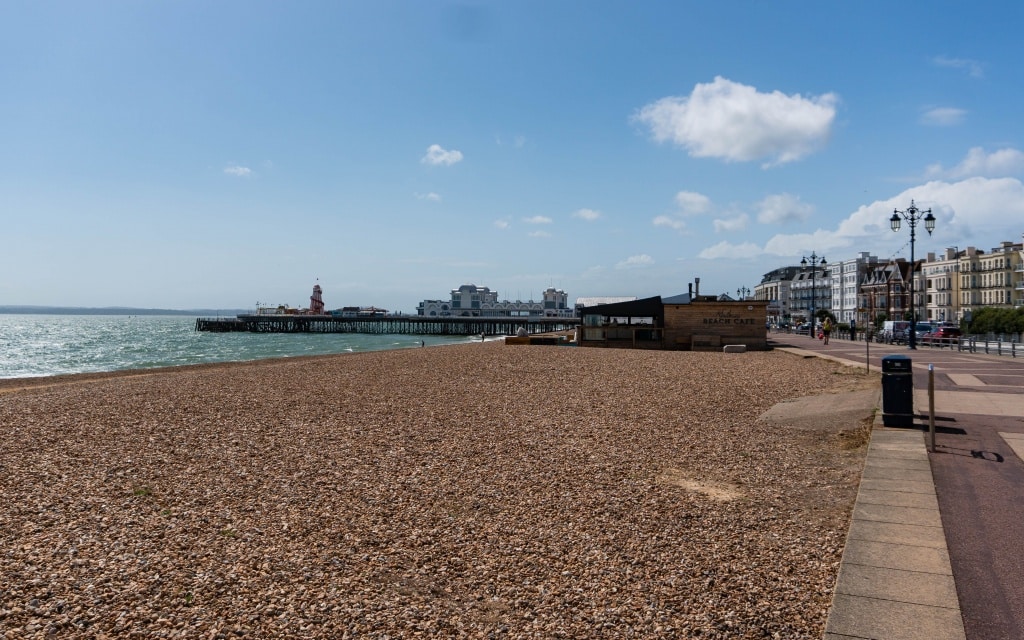 A pebble beach in Southsea features South Parade Pier extending into the sea, lined with buildings and a promenade beneath a sunny blue sky with scattered clouds, near the lively Kidz Island.