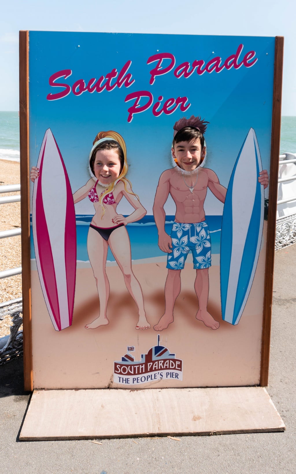 Two people pose with their faces in a seaside cut-out board at South Parade Pier, appearing as a woman in a bikini holding a pink surfboard and a man in swimming trunks with a blue surfboard, with the Southsea beach scene in the background.