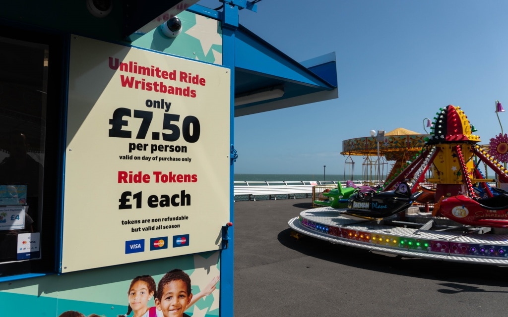 A sign at South Parade Pier in Southsea advertises unlimited ride wristbands for £7.50 per person and ride tokens for £1 each. In the background, Kidz Island rides and the seafront are visible on a sunny day.