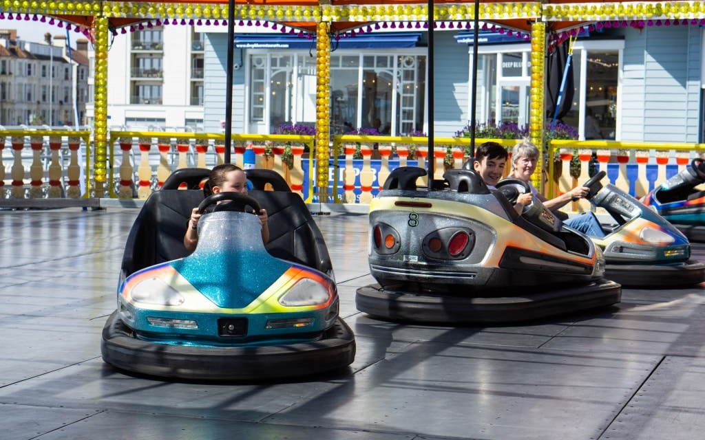 Three children ride dodgems at Kidz Island in Southsea, smiling and steering on a shiny black floor with colourful lights glowing and the South Parade Pier visible against the cityscape in the background.